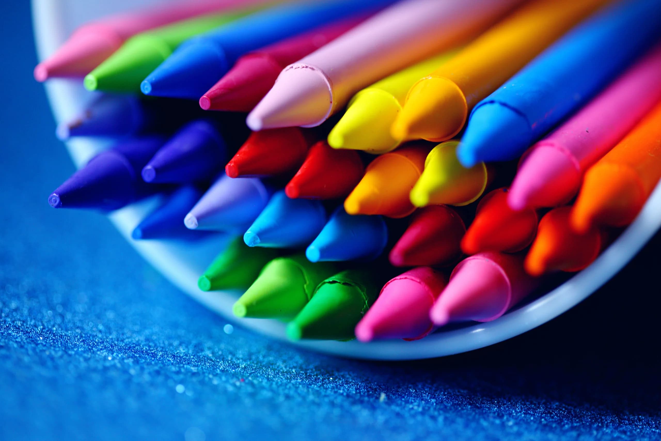 A close-up of vibrant colored crayons arranged in a bowl on a blue surface.